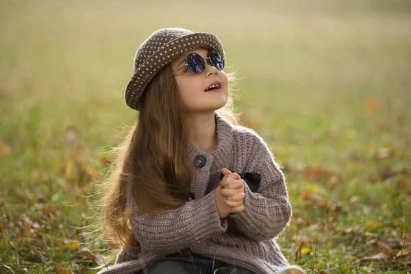 Little girl with sunglasses and hat sitting on a grass in autumn clothes — Stock Photo, Image