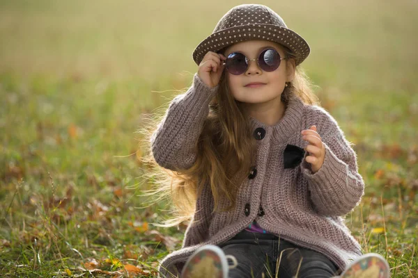 Little girl with sunglasses and hat sitting on a grass in autumn clothes — Stock Photo, Image