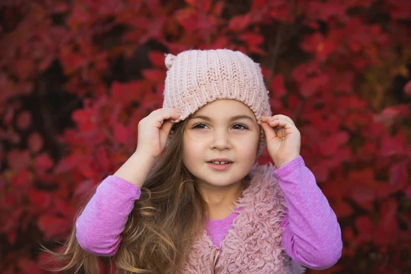 Little girl wearing hat in autumn clothes against red tree foliage background — Stock Photo, Image