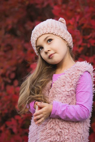 Little girl portrait in hat at autumn clothes against red tree foliage background, she looks up with a cunning look — Stock Photo, Image