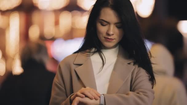 Женщина смотрит время на часы в ночном городе, замедленной съемки, она ждет своего друга — стоковое видео