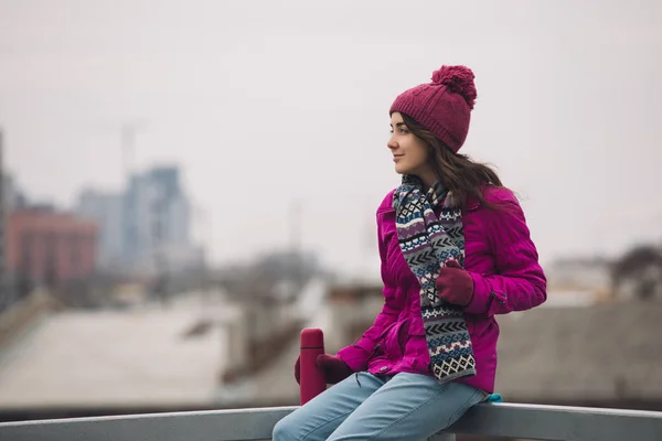 Mujer en chaqueta de invierno brillante sosteniendo termos con té en el fondo de la ciudad —  Fotos de Stock