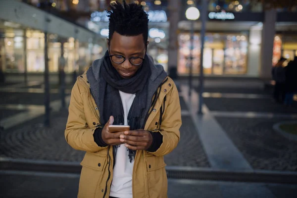 Afrikai ember használ smartphone éjszaka állt sötét város, ő típusú sms üzenet — Stock Fotó