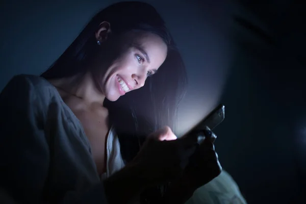 Улыбающаяся женщина с помощью смартфона ночью в темной комнате, сервис онлайн знакомств — стоковое фото