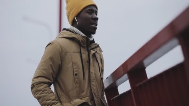 Молодой черный человек холодной зимой слушает музыку на мосту — стоковое видео