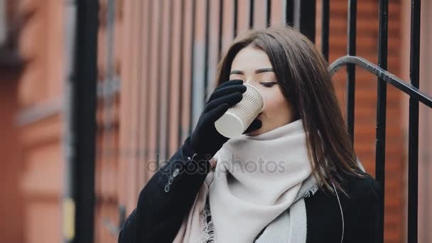 Женщина пьет кофе из бумажной чашки возле забора в холодную зиму — стоковое видео