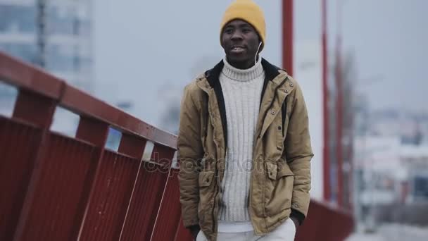 Joven hombre negro en invierno frío escuchando música caminando en un puente — Vídeo de stock