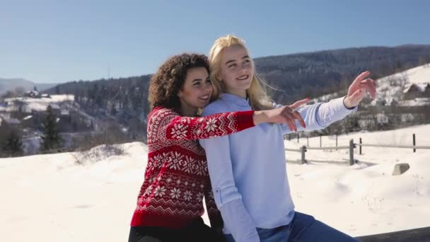 Happy girls friends sitting on fence, showing something together at snowy landscape — Stock Video