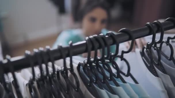 Женщина выбирает одежду в магазине, фокусируется на вешалках — стоковое видео