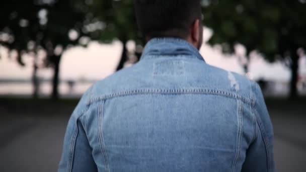 Back of man in torn jeans jacket walking in city park — Stock Video