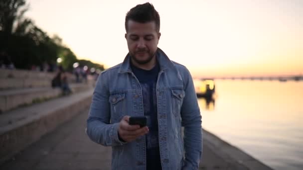 Hombre sonriente usando teléfono inteligente caminando en el terraplén de la ciudad — Vídeo de stock