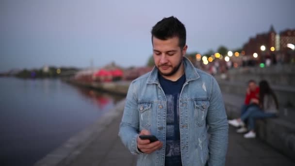 Hombre sonriente usando teléfono inteligente caminando en el terraplén de la ciudad — Vídeos de Stock