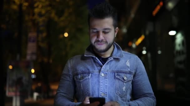 Uomo sorridente utilizzando smartphone a piedi attraverso la strada della città di notte — Video Stock