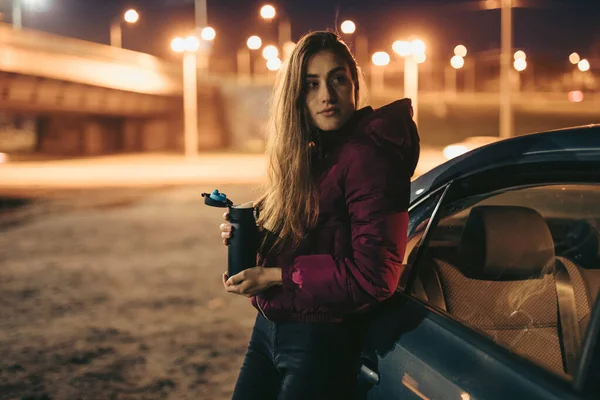 Retrato de mulher motorista segurando garrafa térmica inclinado no carro, tempo da noite — Fotografia de Stock