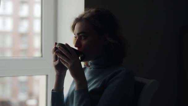 Söt ung kvinna dricker te hemma tittar genom fönstret — Stockvideo