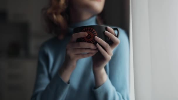 Красивая молодая женщина пьет чай дома, глядя в окно — стоковое видео