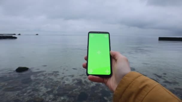 Мужская рука держит чистый зеленый экран смартфона у моря — стоковое видео