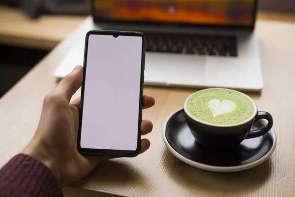 Workplace with matcha latte, male hand holding smartphone with blank screen — ストック写真