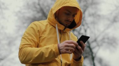 Sarı ceketli ve kapüşonlu bir adam parkta akıllı telefon kullanıyor.