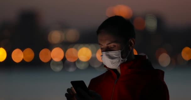 Uomo che utilizza smartphone con maschera chirurgica in una città serale — Video Stock