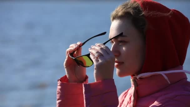 Mulher coloca óculos de sol de pé ao lado de um corpo de água — Vídeo de Stock