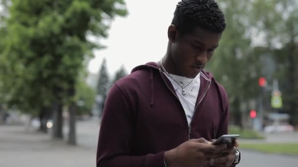Afrika kökenli Amerikalı bir adam yaz şehrinde yürürken akıllı telefona bakar. — Stok video