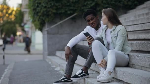 Dos amigos multirraciales hablando sentados en una ciudad — Vídeos de Stock