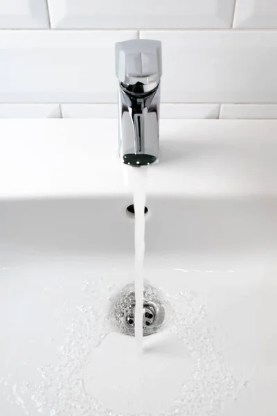 A água flui da torneira para a pia de cerâmica branca e azulejo branco — Fotografia de Stock