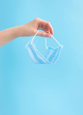 Fotokopi alanı olan mavi arka planda tek kullanımlık tıbbi koruyucu yüz maskesi tutan kadın eli. Koronavirüs salgını, dikey