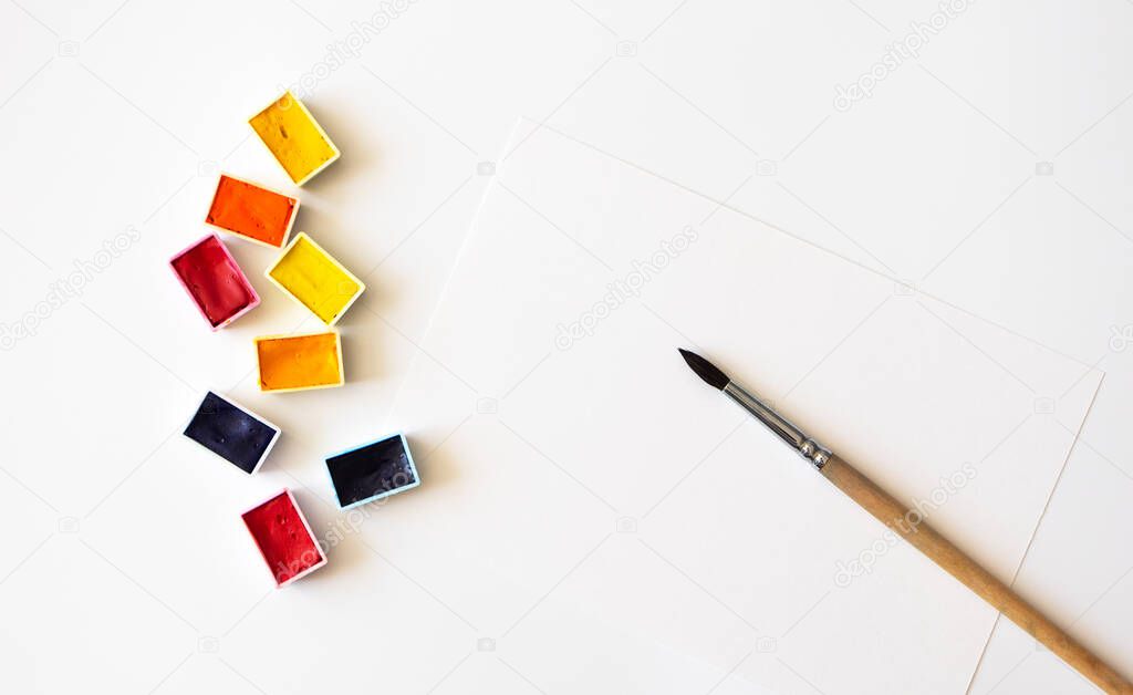 Colorful watercolor pallets, paper sheet and paintbrush on white background, close-up. Hobby and craft, above. Pastime at home