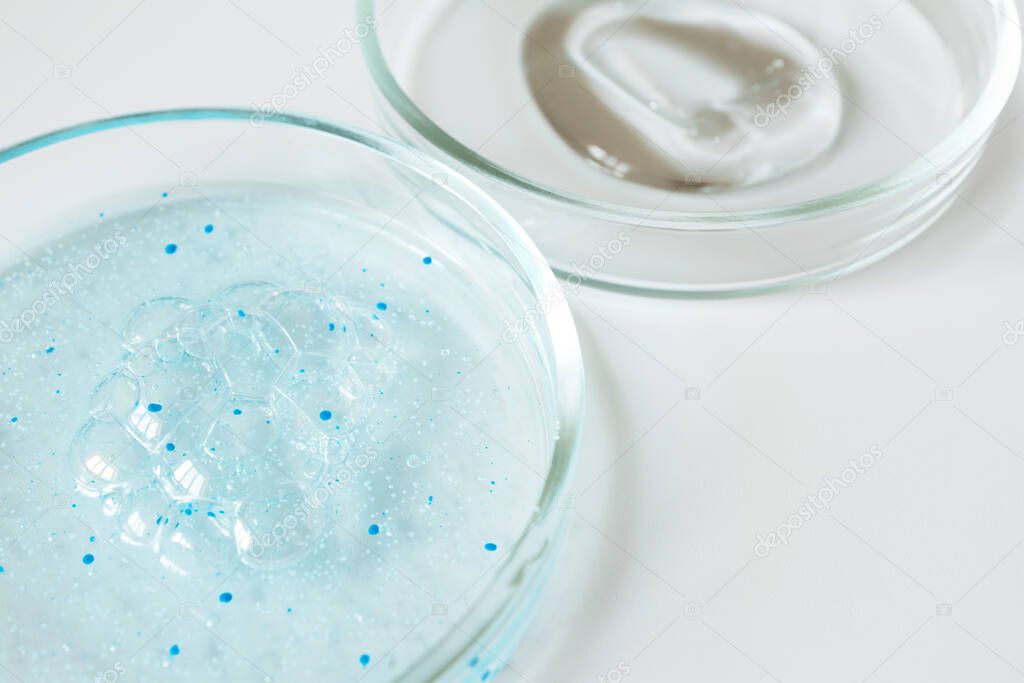 Close-up blue shower gel with scrub grain with bubbles and transparent shampoo smudge in glass petri dish with selective focus on white background. Concept laboratory tests and research