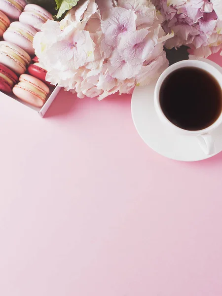 Box with macaroons and cup of coffee, flowers on pink background, copy space, top view
