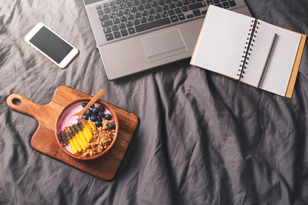 Workplace in bed with laptop and smoothie bowl on grey