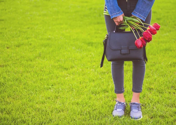 Молодая девушка в джинсах с букетом тюльпанов цветы и сумки прогулки в парке — стоковое фото