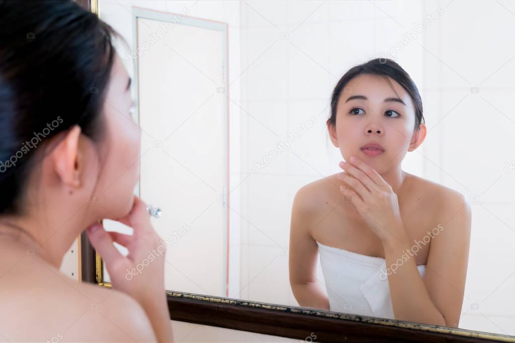 Asian woman squeezing pimples on her face while looking at refle