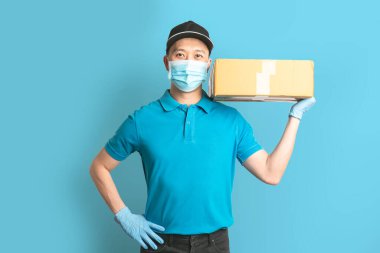 Mavi gömlekli, maske eldivenli teslimatçı eleman mavi arka planda boş karton kutu tutuyor. Coronavirus virüsü 2019 konsepti