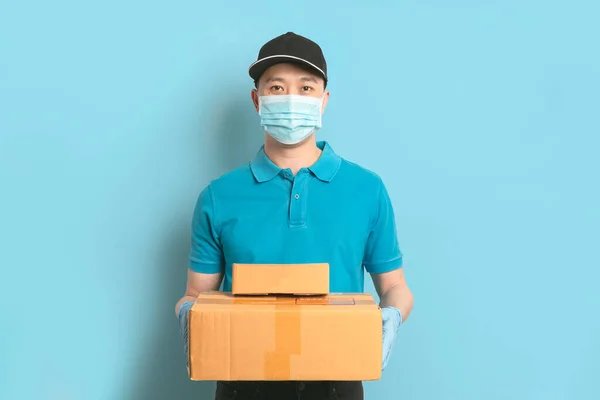 Delivery man employee in blue shirt uniform face mask gloves hold empty cardboard box on blue background. Coronavirus virus 2019 concept