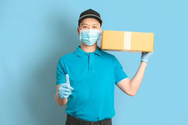 Delivery man employee in blue shirt uniform face mask gloves hold empty cardboard box on blue background. Coronavirus virus 2019 concept