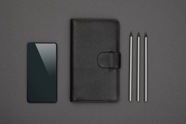 Kapalı siyah deri kapaklı bir defter, kalemler ve akıllı telefonları gri kağıdın üstüne, düz bir zemine, tam üstüne.
