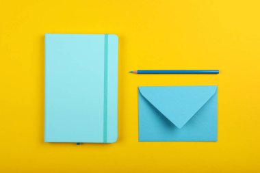 Pastel mavi defterin düzgünce düzenlenmiş kırtasiye düz yatağı, sarı kağıt zemin üzerinde zarf ve kalem, yüksek üst görünüm, tam üzerinde