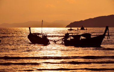 Güneş batan deniz kıyısı, uzun kuyruklu tekne siluetleri ve parlak gökyüzü üzerinde ufuk çizgileri.