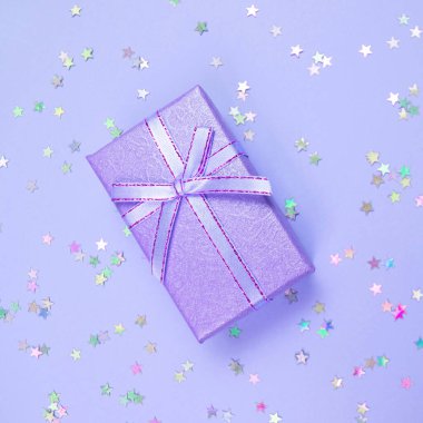 Proton Mor hediye kutusu, kurdele ve gökkuşağı holografik yıldızları menekşe pastel arka planda konfeti.