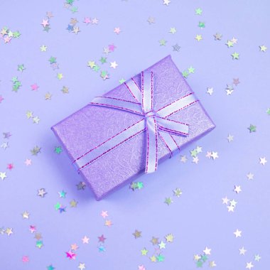 Proton Mor hediye kutusu, kurdele ve gökkuşağı holografik yıldızları menekşe pastel arka planda konfeti.