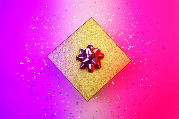 Caja de regalo dorada con cinta y estrellas holográficas del arco iris confeti. Mínimo plano yacía en luz de neón led — Foto de Stock