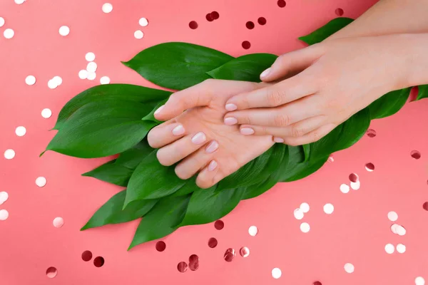 Due mani di donne su foglie verdi. Nudo manicure gel femminile su sfondo rosso corallo con coriandoli scintillanti. Posa piatta — Foto Stock
