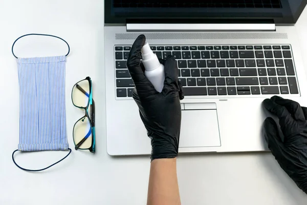 Two women\'s hands in black latex gloves disinfect a computer with an antibacterial spray. Office gadget disinfection at work top view. Laptop, medical mask, antiseptic virus protection health care