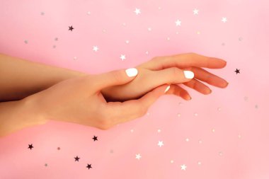 Pembe arka planda kusursuz beyaz manikürlü, yıldız konfetiyle iki hassas kadın eli. Üstten yakından bak. Güzellik, moda, kaplıca salonu düz konsept. Moda sağlık pastel renk tırnak cilası.