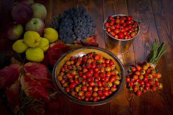 Clay bowl full of rose hips on wooden background. Autumn fruits in the background. — Stock Photo, Image