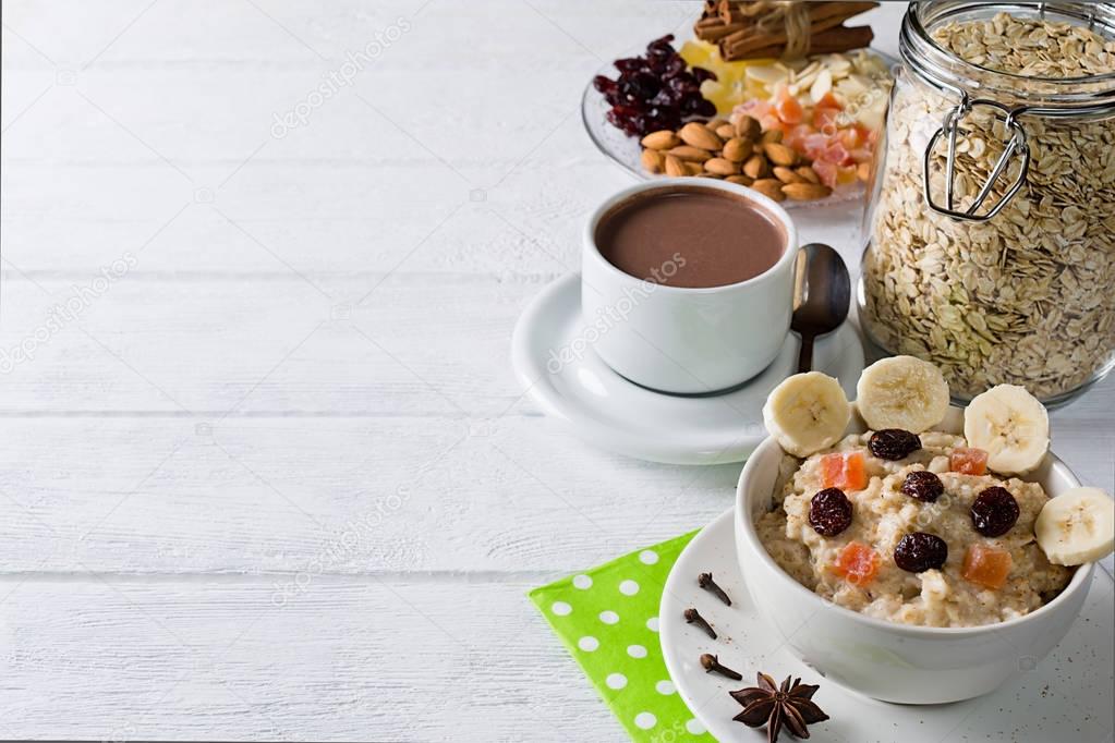 Oatmeal porridge with dried fruits, cranberries, bananas and spices. Cup of cacao and jar of oatmeal on background. Arranged on white background.