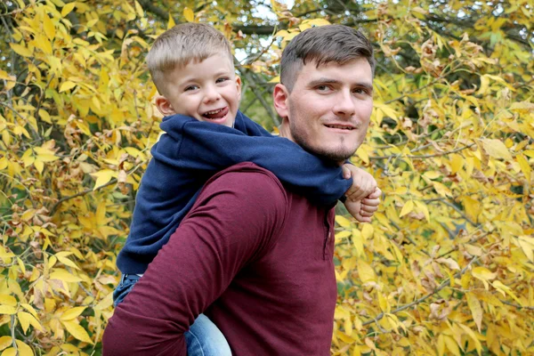 El padre fuerte sostiene a su pequeño hijo guapo lindo en su espalda al aire libre en el bosque —  Fotos de Stock